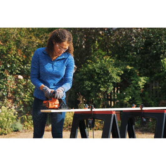Person using BLACKDECKER BDCCS18N cordless circular saw to cut wood outdoors, with workpiece clamped to sawhorses.