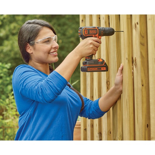 Person using BLACKDECKER BDCDC18 cordless drill to drive screw into vertical wooden planks outdoors.