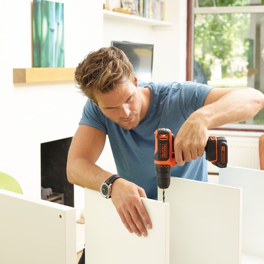 BLACKDECKER BDCDD12A80T cordless drill in use, person drilling into white furniture panel indoors.