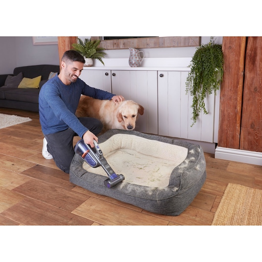BLACKDECKER BHHV520BFP handheld vacuum cleaning pet hair from a gray dog bed, person kneeling beside golden retriever.